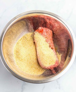 A stainless steel bowl containing raw ground meat partially covered with breadcrumbs on a marble countertop.