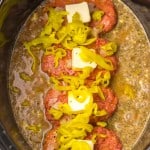 Raw burger patties topped with butter and sliced peppers in a slow cooker, surrounded by liquid and diced onions.