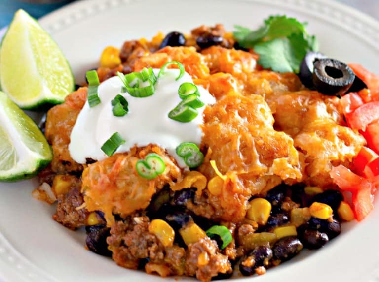 Plate of nachos with ground beef, black beans, corn, melted cheese, sour cream, chopped green onions, olives, and diced tomatoes, garnished with lime wedges and cilantro.