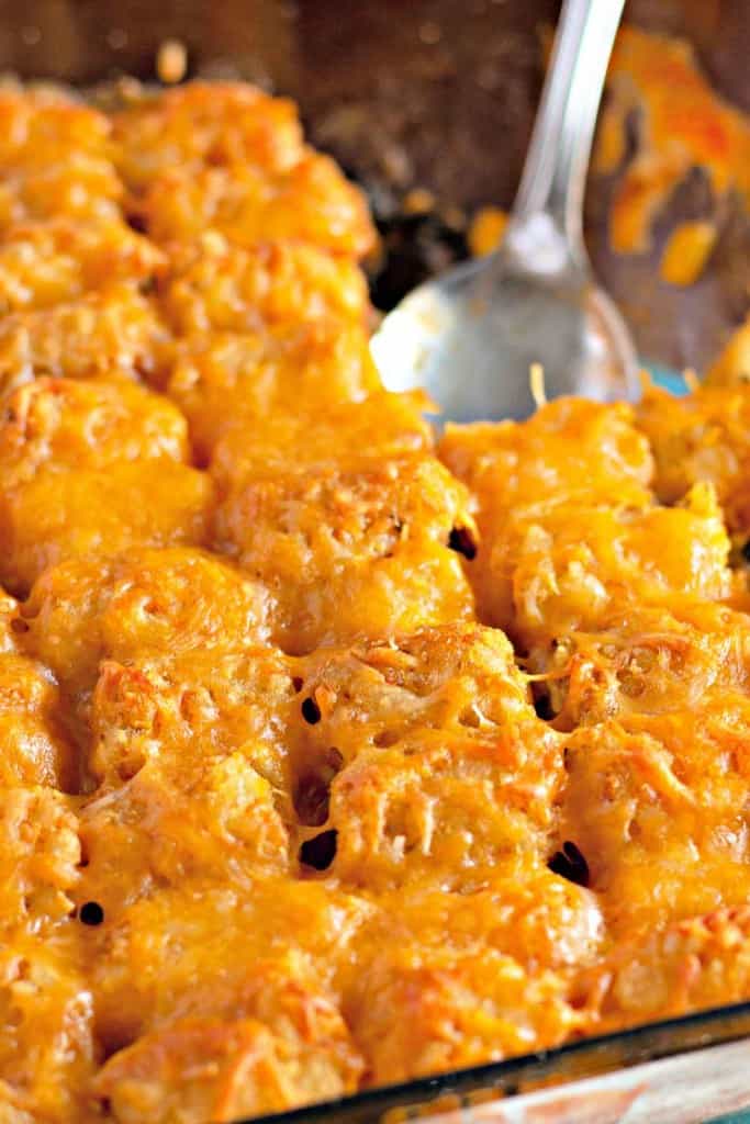A casserole dish filled with baked tater tots covered in melted cheese, with a serving spoon on the side.