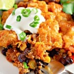 A plate of taco casserole with beans, corn, ground meat, and melted cheese, topped with sour cream and chopped green onions, garnished with a lime wedge and cilantro.
