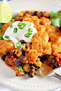 A plate of taco casserole with beans, corn, ground meat, and melted cheese, topped with sour cream and chopped green onions, garnished with a lime wedge and cilantro.
