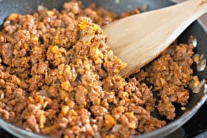 Cooked ground beef with onions in a skillet, being stirred with a wooden spatula.