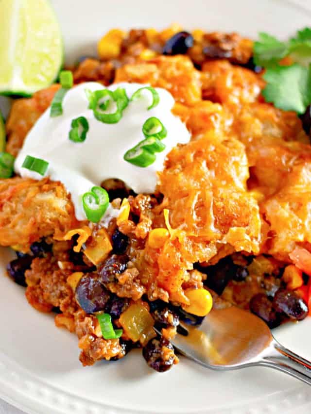 A plate of taco casserole topped with sour cream and chopped green onions, with a fork beside it, garnished with a lime wedge and cilantro.