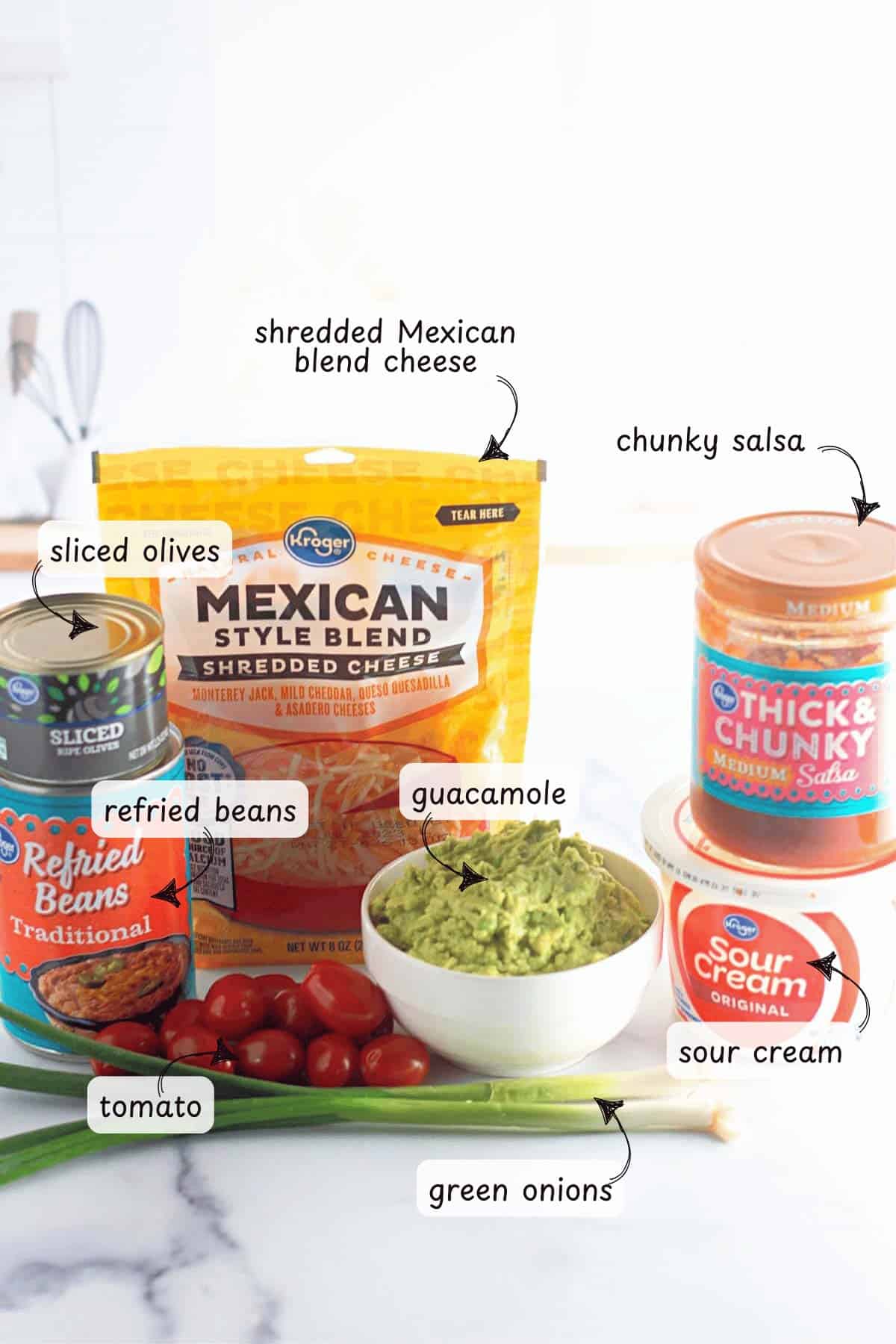 Ingredients for 7 layer dip cups are arranged on a table, featuring shredded cheese, guacamole, sour cream, salsa, refried beans, sliced olives, tomatoes, and green onions.