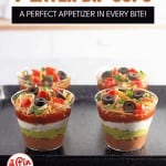 Individual cups of seven-layer dip arranged on a tray, each topped with shredded cheese, sliced olives, and diced tomatoes.