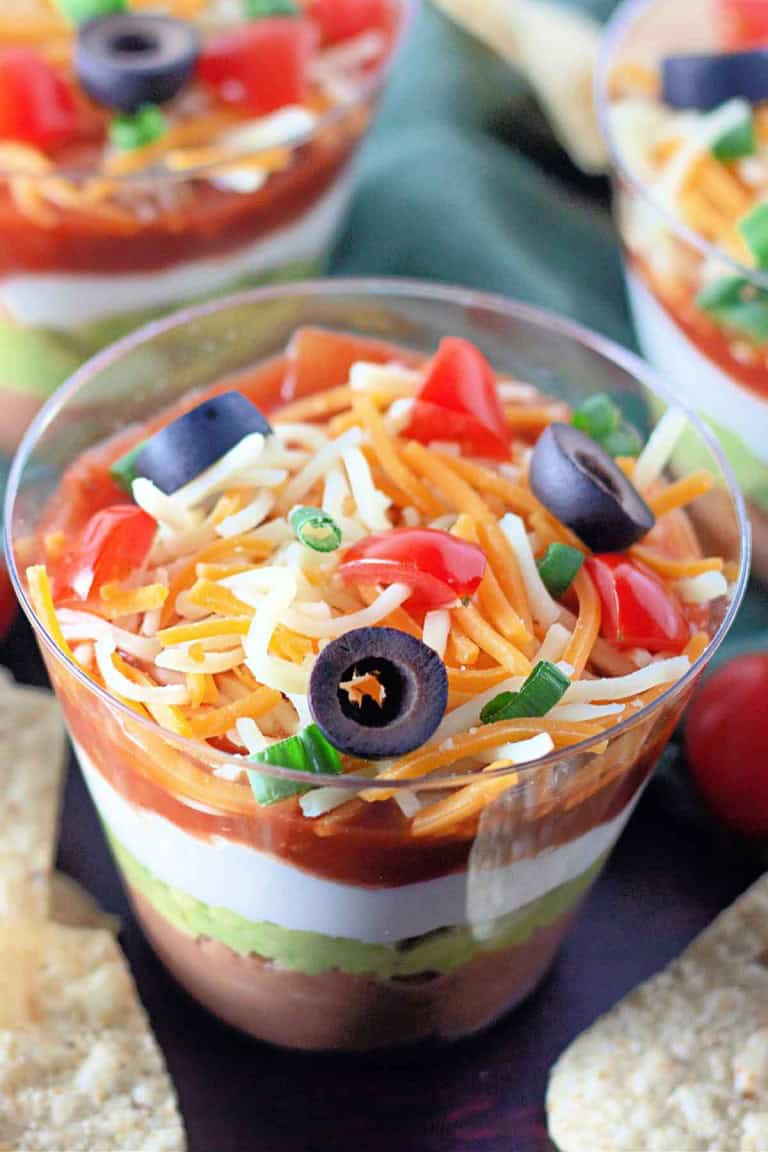 Close-up of a seven-layer dip in a clear cup, featuring layers of beans, guacamole, sour cream, salsa, cheese, and toppings of chopped tomatoes, black olives, and green onions.