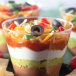 A layered dip in a cup featuring beans, guacamole, sour cream, salsa, shredded cheese, olives, and green onions.