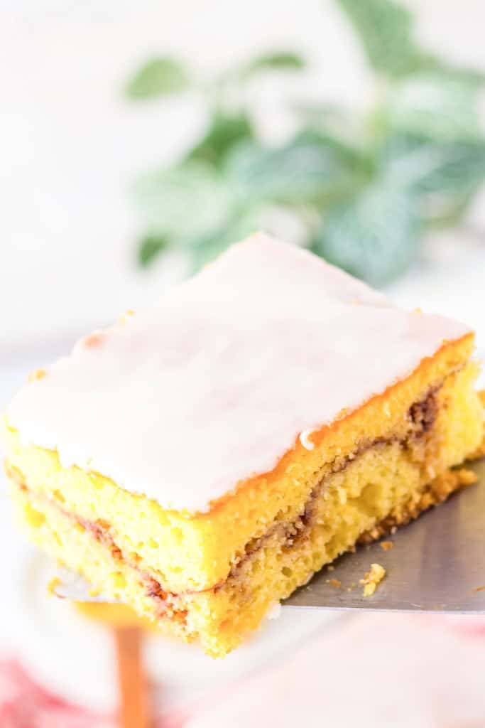 A piece of frosted yellow cake with visible cinnamon swirls on a spatula, set against a blurred background with green leaves.