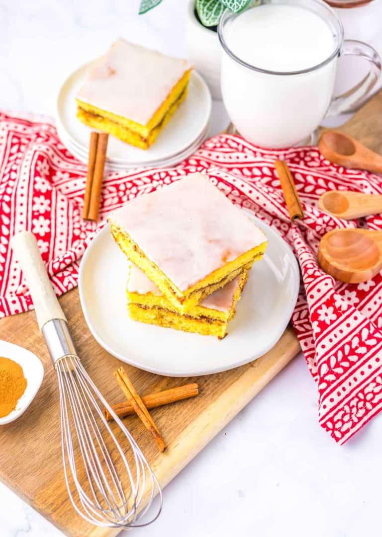 Glazed dessert bars on plates, surrounded by cinnamon sticks, a whisk, wooden spoons, a red patterned cloth, and a cup of milk on a wooden board.