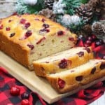 Cranberry bread on a wooden board, with slices cut, surrounded by cranberries on a red and black checkered cloth, highlights the vibrant flavors of cranberry recipes this December. Pinecones linger in the background, adding a touch of seasonal charm.