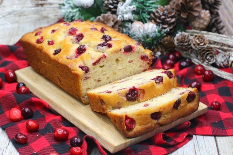 Cranberry bread on a wooden board, with slices cut, surrounded by cranberries on a red and black checkered cloth, highlights the vibrant flavors of cranberry recipes this December. Pinecones linger in the background, adding a touch of seasonal charm.