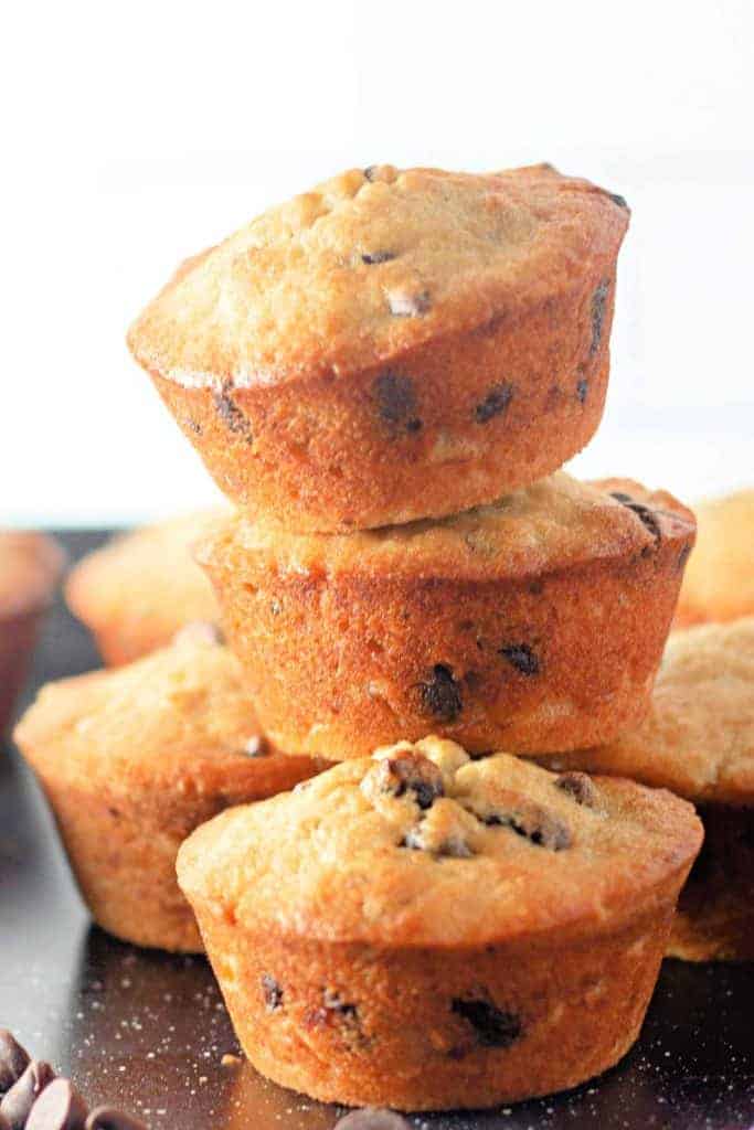A stack of three chocolate chip muffins on a dark surface, with more muffins blurred in the background.