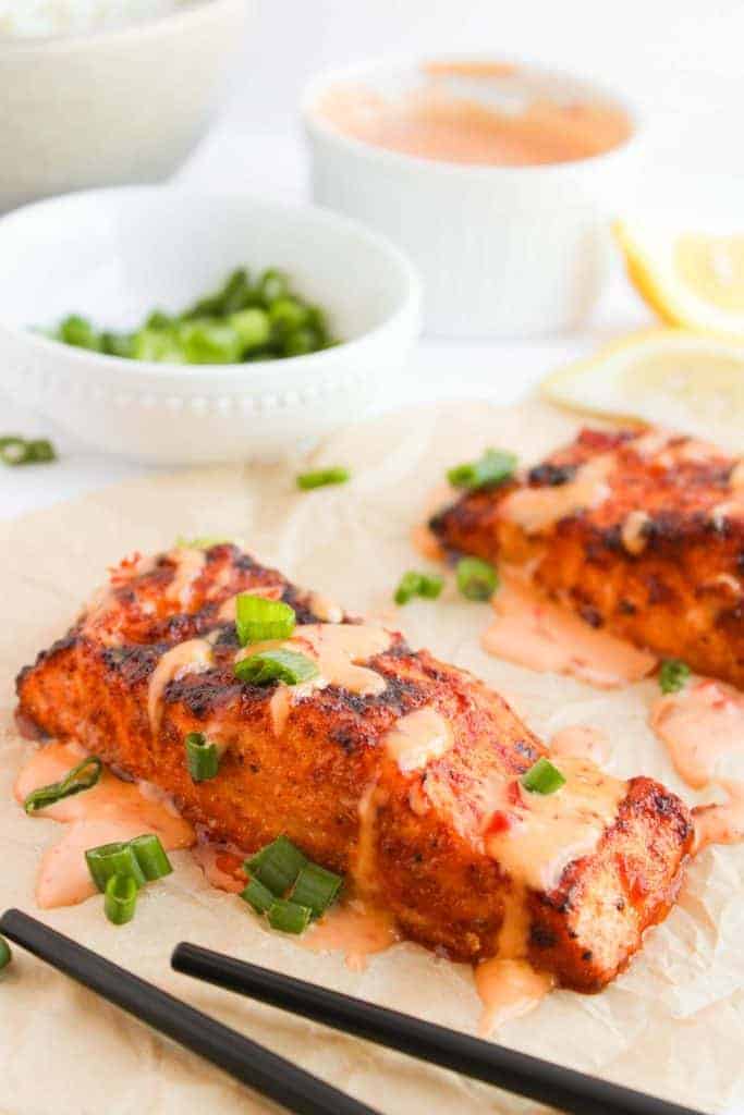 Two grilled salmon fillets topped with sauce and green onions, placed on parchment paper. Black chopsticks and a bowl of green onions are nearby.