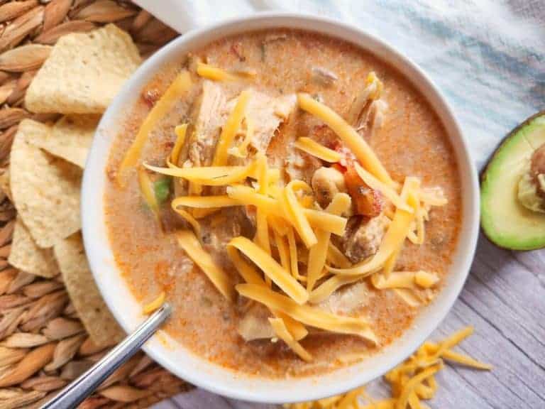A bowl of creamy soup topped with shredded cheese, served with tortilla chips and a sliced avocado on the side.