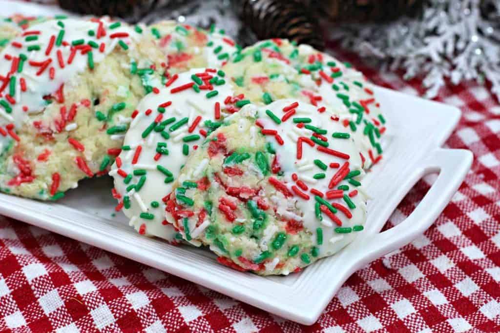 Three cookies with white icing and red, green, and white sprinkles on a rectangular white plate. The plate is on a red and white checkered tablecloth.