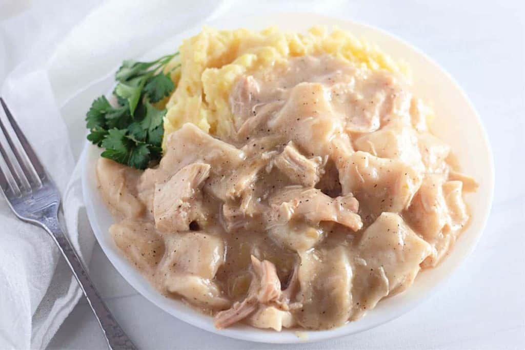 A hearty plate of creamy chicken and dumplings, reminiscent of classic stews, paired with mashed potatoes and garnished with cilantro, sits invitingly next to a fork.