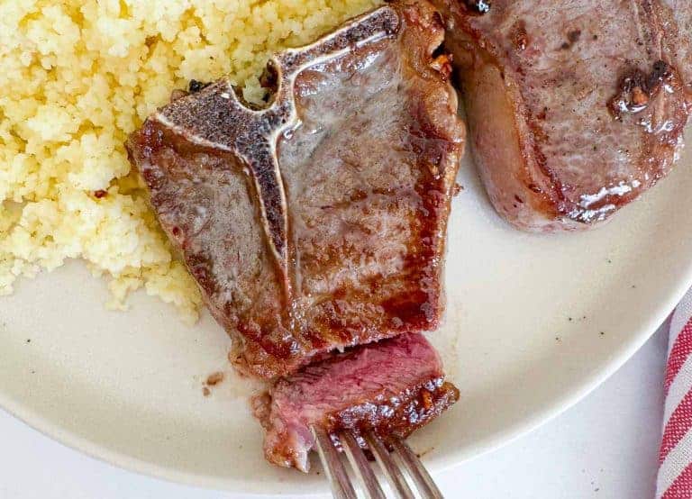 A plate with cooked lamb chops, one partially cut, served next to couscous. A fork holds a piece of lamb chop.