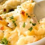 A bowl of mashed potatoes topped with chopped parsley, with a spoon scooping a portion.