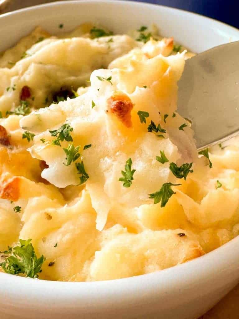 A bowl of mashed potatoes topped with chopped parsley, with a spoon scooping a portion.