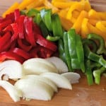 Sliced red, green, and yellow bell peppers with white onion on a wooden cutting board create the perfect base for a hearty Kielbasa Skillet.