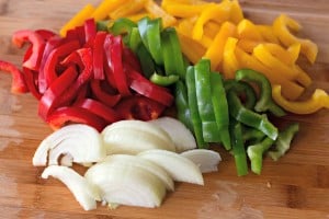 Sliced red, green, and yellow bell peppers with white onion on a wooden cutting board create the perfect base for a hearty Kielbasa Skillet.