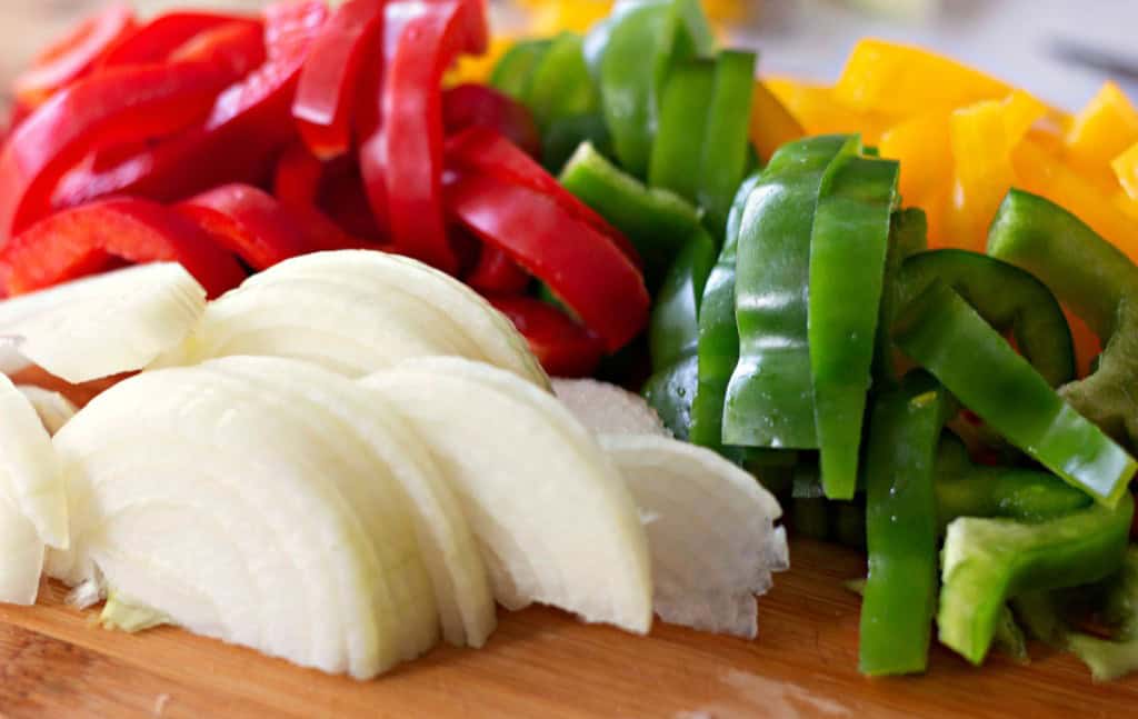 Sliced vegetables on a wooden board, featuring red and yellow bell peppers, green bell peppers, and white onions—perfect for adding vibrant flavor to a pierogi and kielbasa skillet.