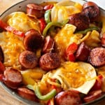 A skillet filled with kielbasa, sliced sausages, red and green bell peppers, onions, and melted cheese on top of sliced potatoes creates a delicious harmony reminiscent of a classic pierogi meal.