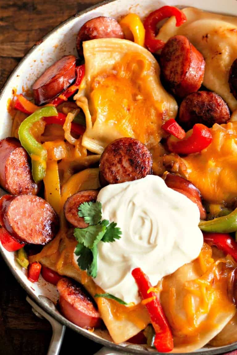 A Pierogi and Kielbasa Skillet featuring sliced sausage, bell peppers, and melted cheese, all topped with a dollop of sour cream and garnished with a cilantro leaf.