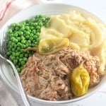 White plate with shredded meat, mashed potatoes, peas, and pepperoncini. A fork is placed on the plate.