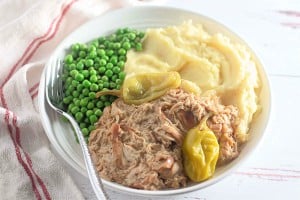 White plate with shredded meat, mashed potatoes, peas, and pepperoncini. A fork is placed on the plate.