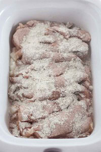 Raw chicken pieces in a white slow cooker, covered with a seasoning mixture.
