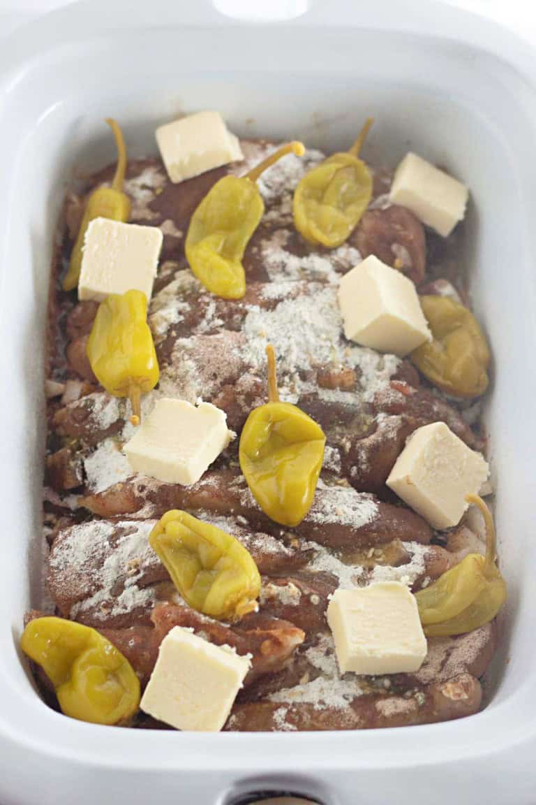 Raw meat in a slow cooker topped with yellow peppers, cubes of butter, and a light sprinkle of seasoning mix.