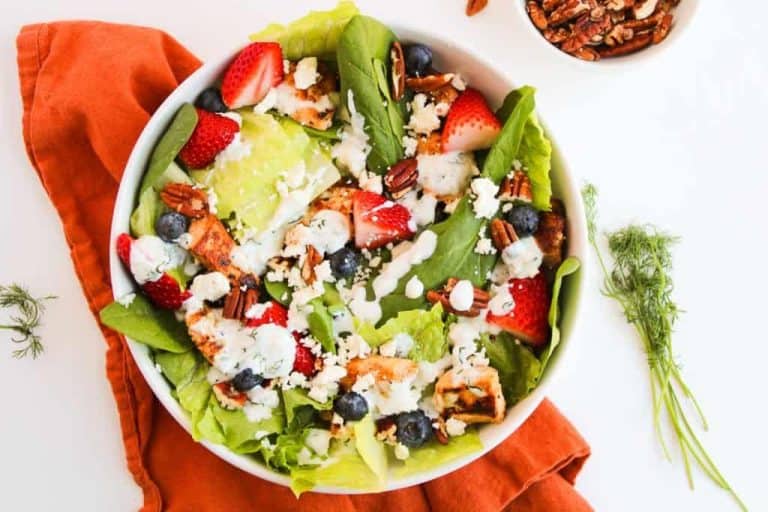 A bowl of salad with lettuce, strawberries, blueberries, grilled chicken, pecans, and crumbled cheese on an orange cloth with a dill garnish nearby.