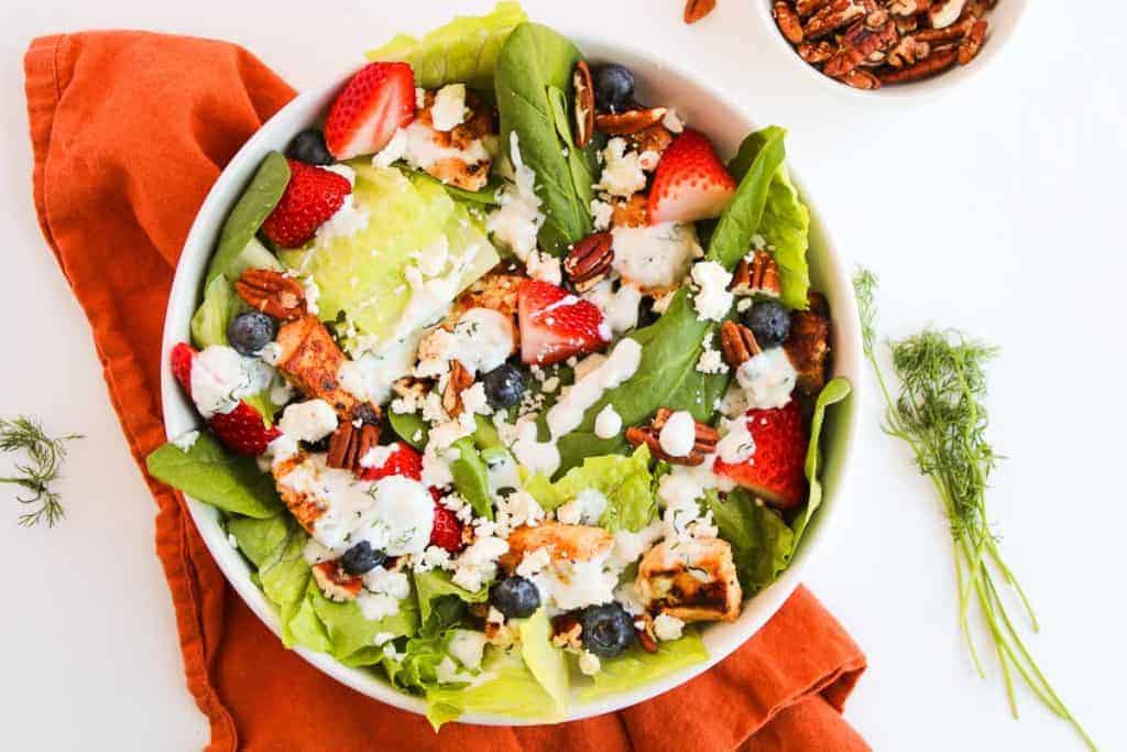 A bowl of salad with lettuce, strawberries, blueberries, grilled chicken, pecans, and crumbled cheese on an orange cloth with a dill garnish nearby.