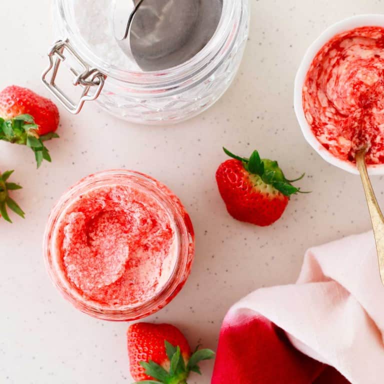 Overhead view of a jar and bowl of strawberry cream with a spoon, surrounded by fresh strawberries and a red napkin on a white surface.