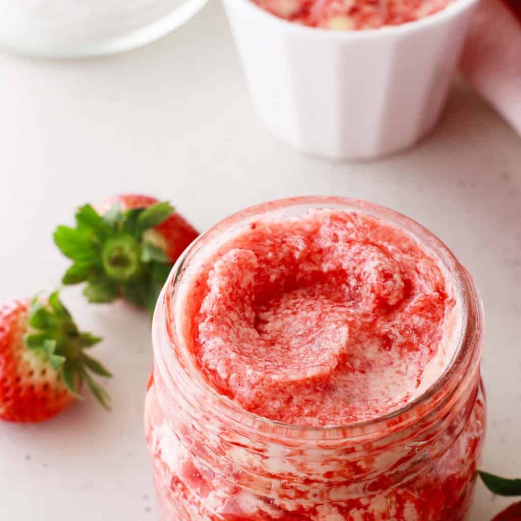 Jar of pink and white swirled strawberry butter on a countertop, with fresh strawberries nearby.