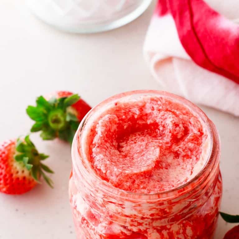 A jar of whipped strawberry butter on a surface, surrounded by fresh strawberries and a red and white cloth.