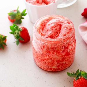 A jar of pink whipped strawberry butter is placed on a countertop, surrounded by fresh strawberries.