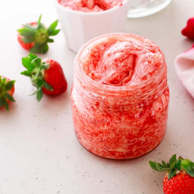 A jar of pink whipped strawberry butter is placed on a countertop, surrounded by fresh strawberries.