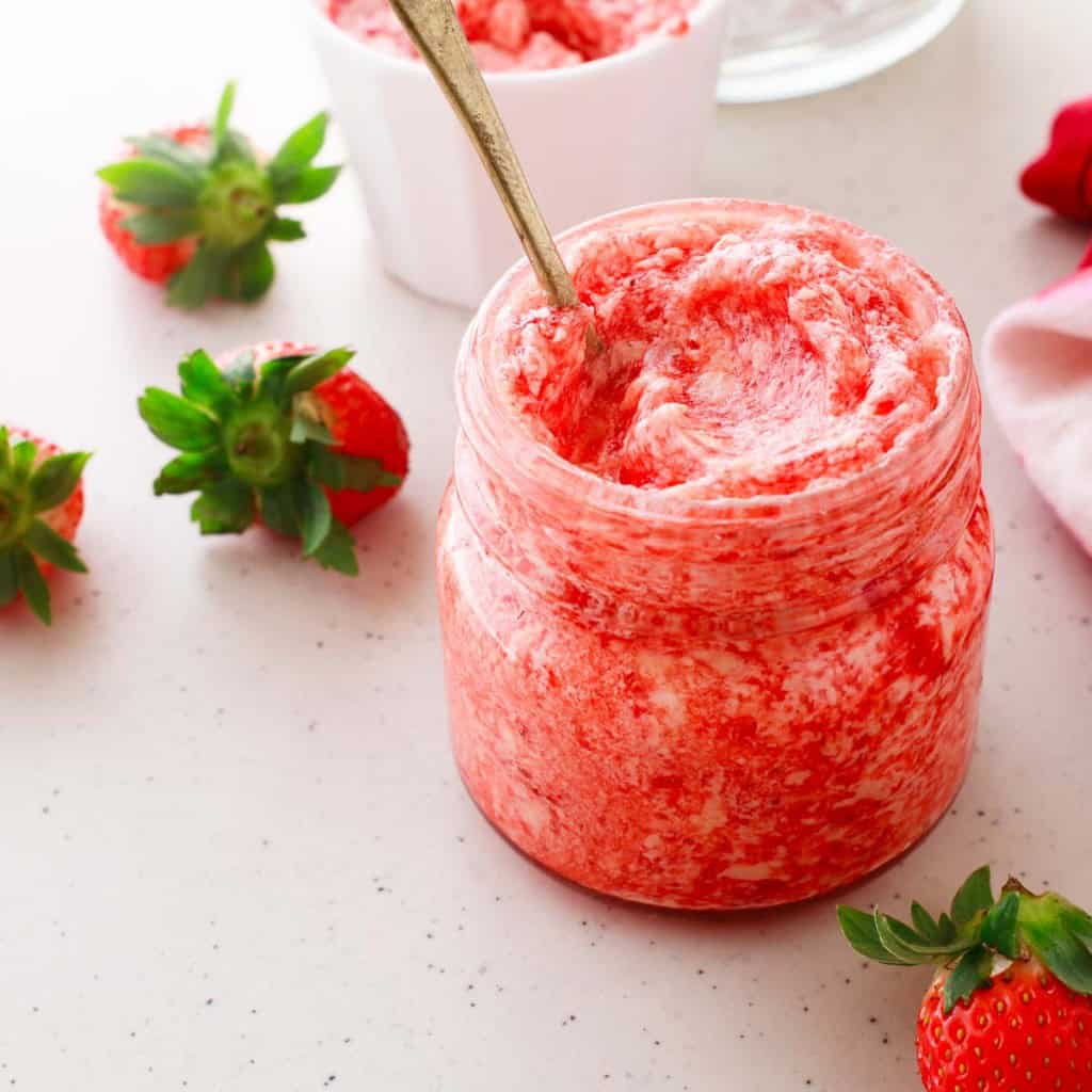 Jar of strawberry butter with a spoon inside, surrounded by fresh strawberries on a light countertop.