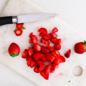 Sliced and whole strawberries on a white cutting board with a knife.