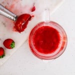 A blender knife next to a few whole strawberries and a clear measuring cup with red pureed strawberries on a white countertop.
