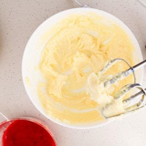 A mixing bowl with creamy yellow batter and a hand mixer. A container with red sauce sits nearby on a light countertop.