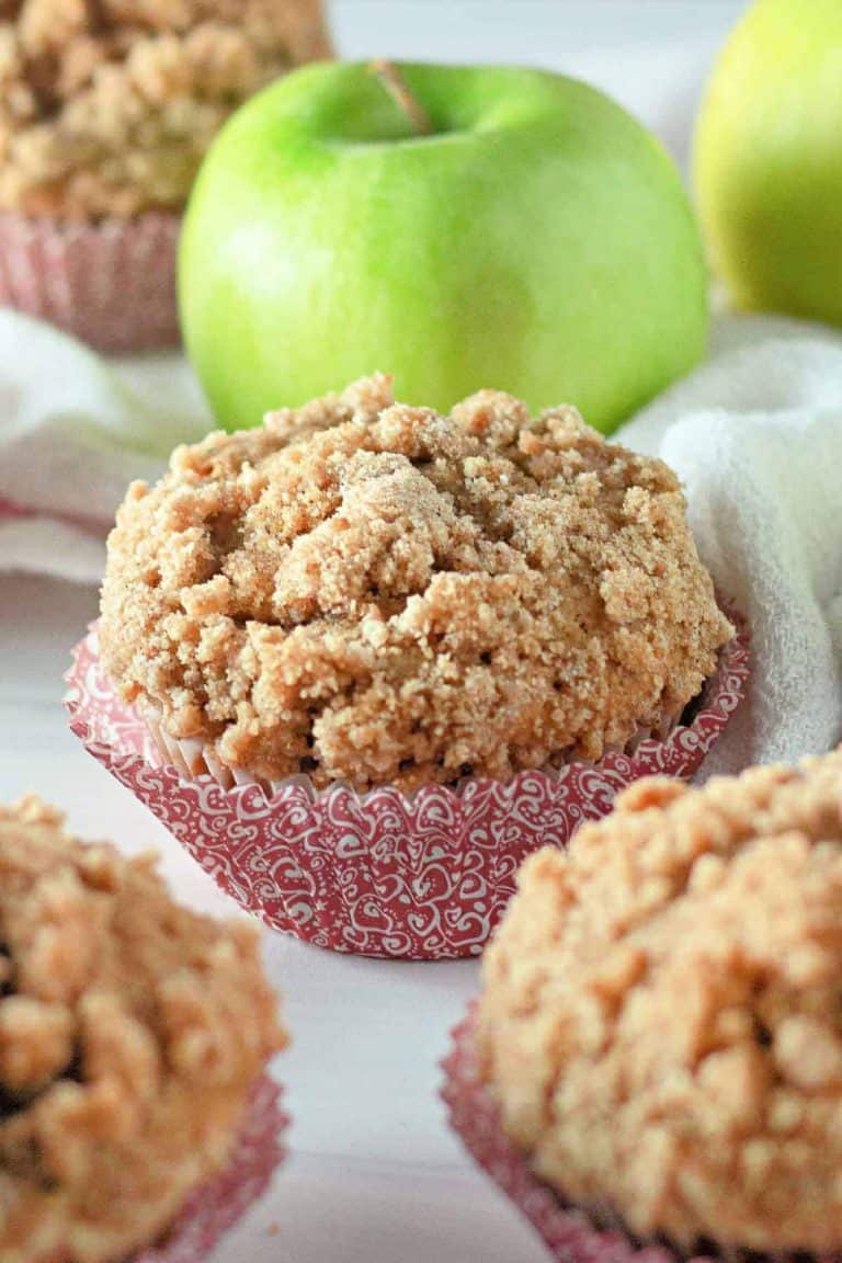 Apple muffins with crumbly topping are displayed in decorative paper cups, surrounded by green apples on a white surface.