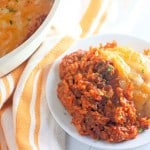 A white plate with a portion of shepherd's pie, featuring a layer of mashed potatoes over seasoned ground meat, is displayed, evoking comfort much like a taco rice skillet. A larger dish of the same meal is partially visible in the background.