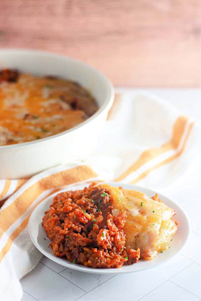 A serving of shepherd's pie with a browned cheese topping on a white plate, reminiscent of a taco rice skillet, next to a larger dish of the same pie, placed on a striped cloth.