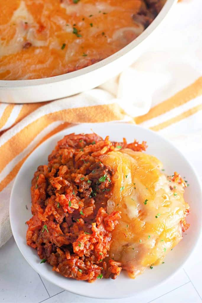 A plate of cheesy taco rice skillet casserole, with ground meat and melted cheese on top, sits next to a baking dish on an orange and white striped cloth.