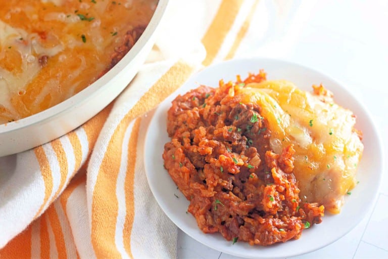 A white plate with cheesy lasagna and a fork on the side, reminiscent of the warmth of a taco rice skillet. A striped towel and a dish of lasagna rest in the background, inviting you to indulge.