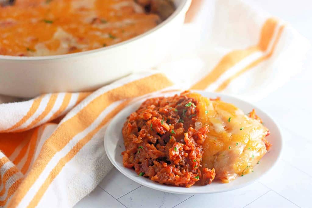 Plate of cheesy lasagna topped with tomato and meat sauce next to a striped napkin, reminiscent of a hearty taco rice skillet. A larger dish is partially visible in the background.